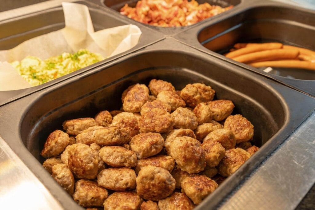 In der Theke des Frühstücksbuffets sieht man im Vordergrund eine große Portion klienre Klopse, im Hintergrund sieht man Rührei, Bacon und Würstchen.